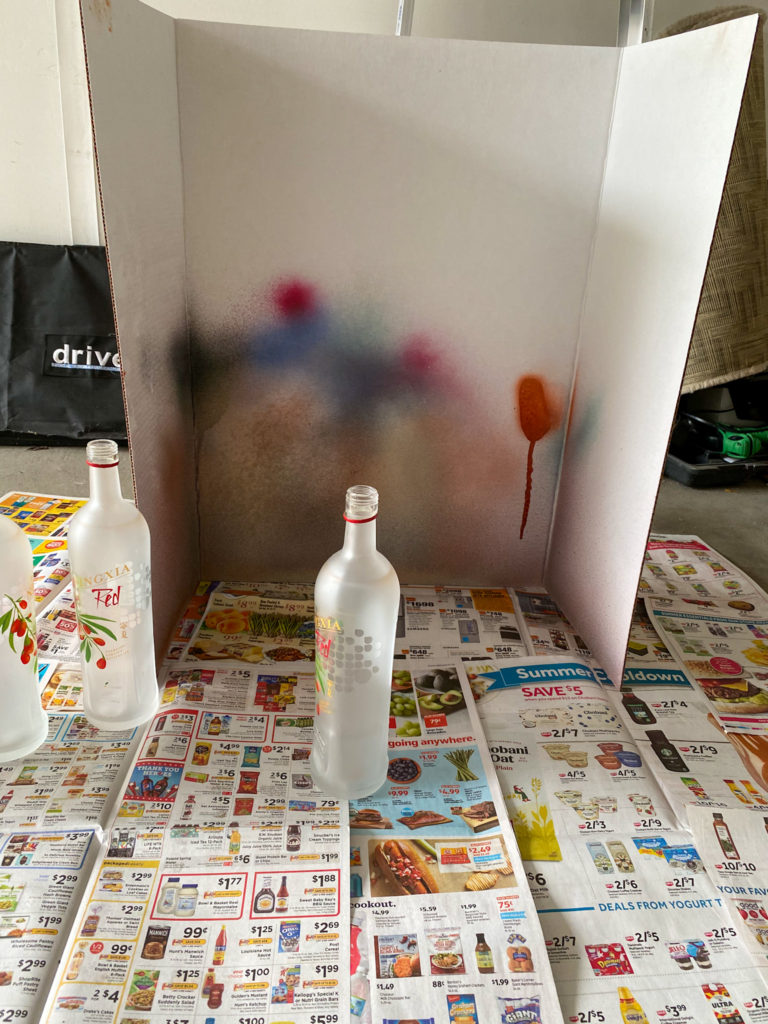 Cardboard trifold with spray paint splotches on top of newspaper with empty glass bottles in front