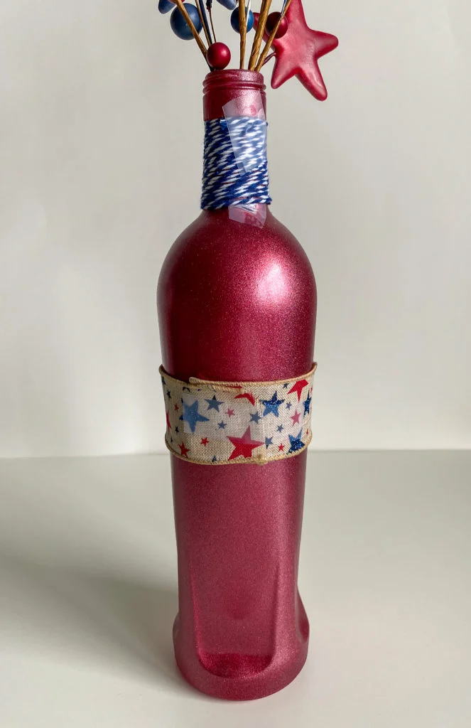 red bottle side view with burlap ribbon with red and blue stars, blue and white twine on neck, red star spray and blue &red berry spray