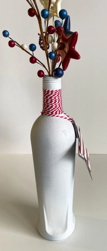 White glitter bottle side view with red & white bakers twine on neck, red/white/blue berry sprays and stars