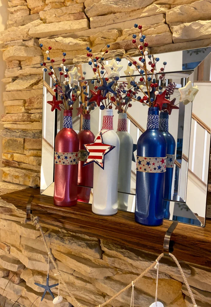 red, white and blue bottles on fireplace mantle in front of mirror, with red, white and blue bakers twine on necks, burlap ribbon with red and blue stars, and sprays of red/white/blue berries and stars