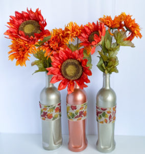 Two caramel latte bottles flanking candy corn bottle, decorated with burlap ribbon with fall leaves design, and filled with rust colored sunflowers and mums