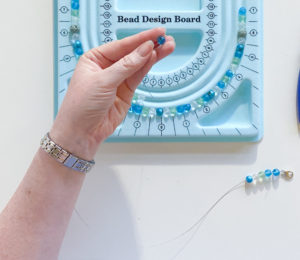 Threading seaglass colored beads onto bead sringing wire, in front of bead desgin board