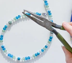 Needlenose pliers holding crimp bead cover, in front of seaglass colored mask chain