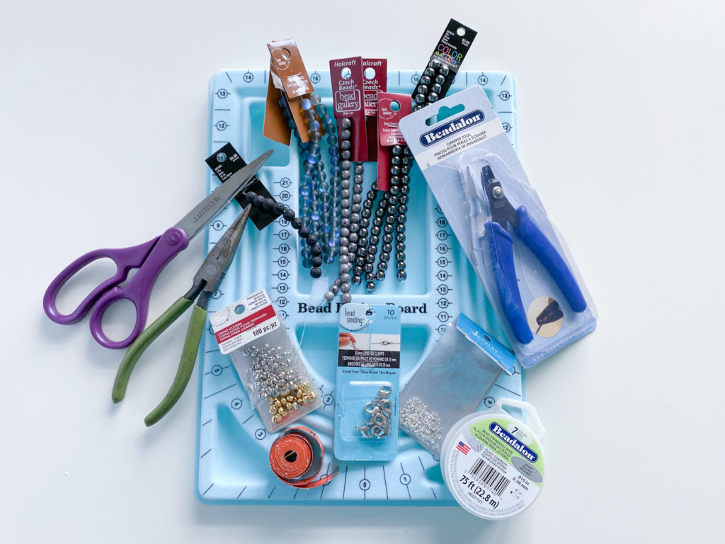 All supplies needed to make gray mask chain: beads, scissors, pliers, crimp beads, crimp covers, lobster clasps, bead stringing wire, crimping tool, measuring tape and bead design board