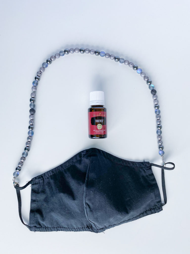 Gray beaded mask chain with black fabric face mask and bottle of Thieves essential oil