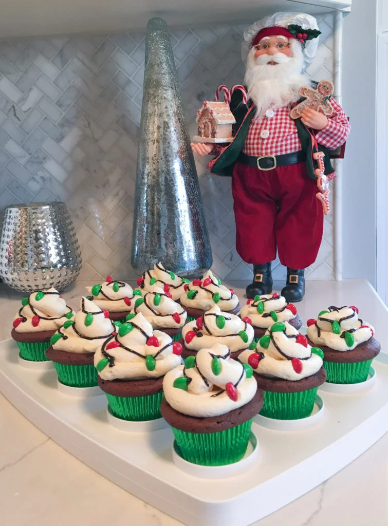 Tray of cupcake carrier with 12 standard sized cupcakes, decorated with red and green M&M Christmas lights, in front of baker Santa