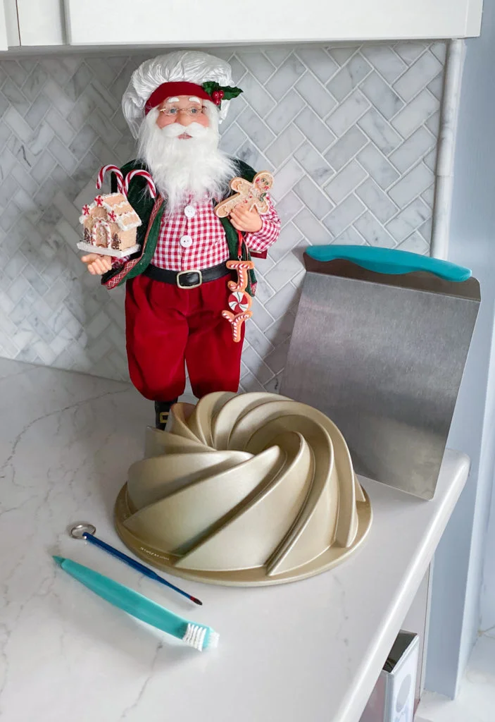 Heritage Bundt pan, Wilton cake lifter, bundt cake thermometer, and bundt cleaning tool in front of baker Santa