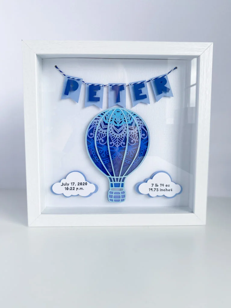 Completed hot air balloon birth announcement shadow box mandala with two clouds containing date and time of birth and vital statistics, and 3D pennant banner spelling out Peter strung with blue and white twine in a white shadowbox frame