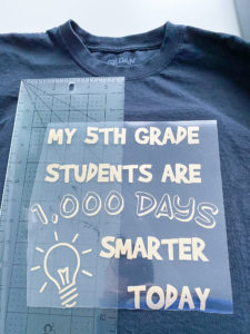 Cricut ruler measuring 3 inches from the bottom edge of center seam of black crew neck t-shirt, laid underneath the weeded image that reads "My 5th Grade Students are 1,000 Days Smarter Today" with lightbulb