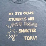 Black t-shirt with gold design reading "My 5th Grade Students are 1,000 Days Smarter Today" with light bulb