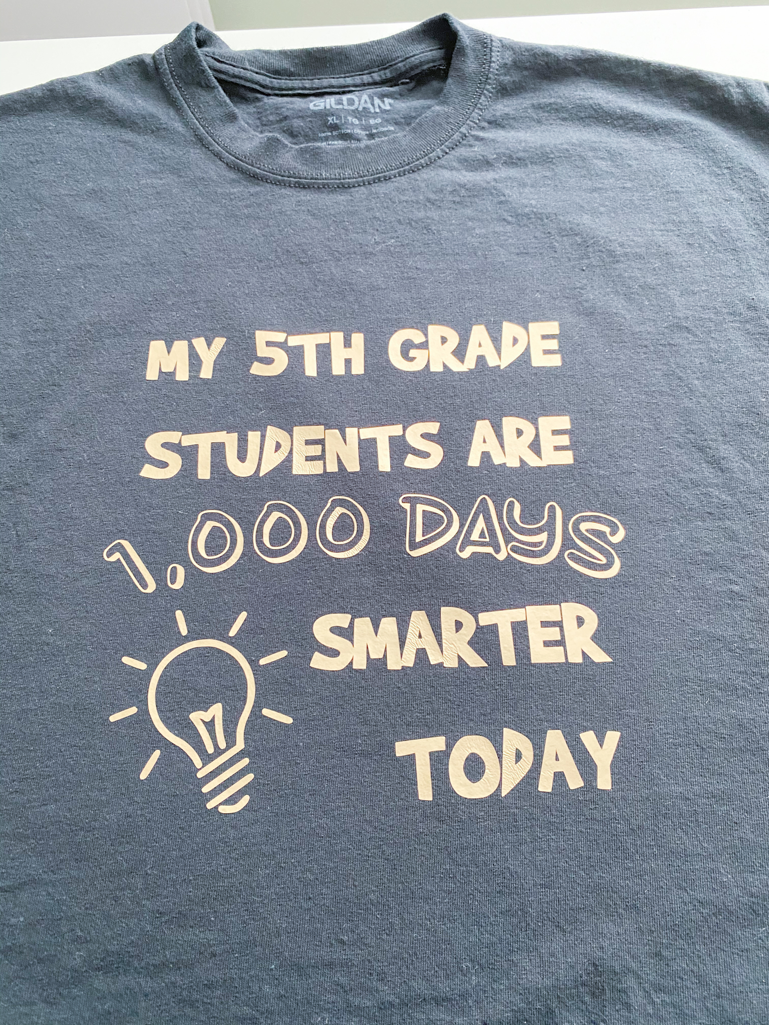 Black t-shirt with gold design reading "My 5th Grade Students are 1,000 Days Smarter Today" with light bulb