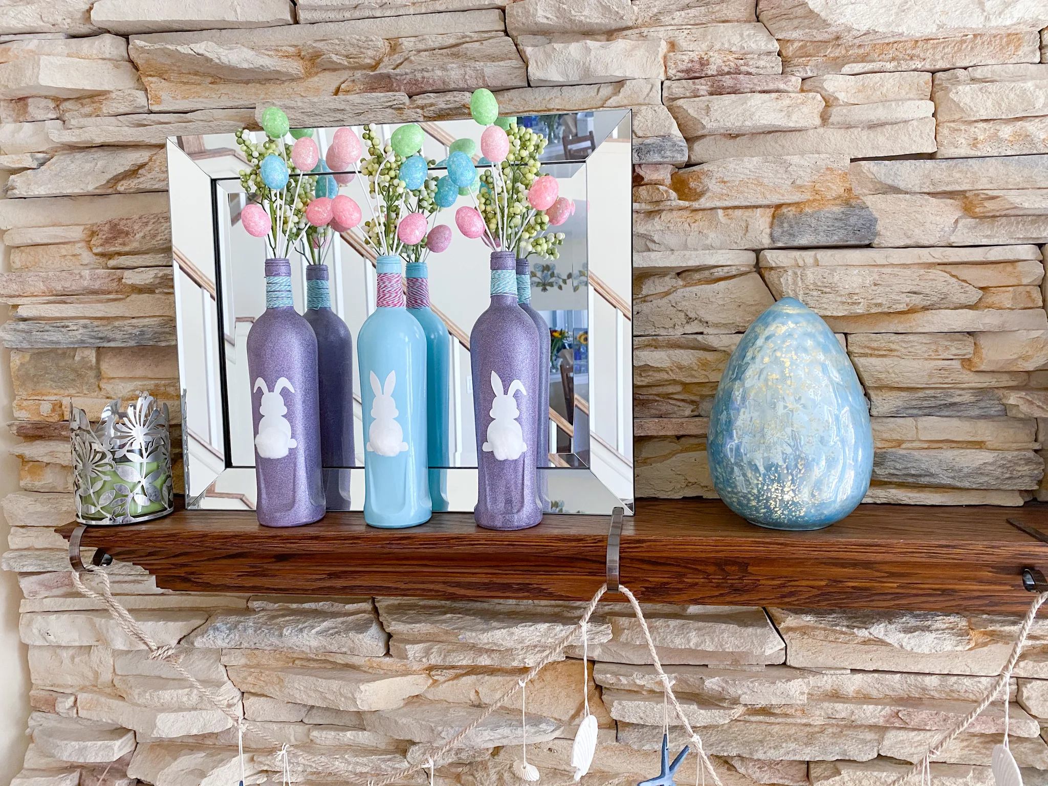 2 purple & 1 aqua bottle with white bunny silhouette labels, cotton ball tails, pink & aqua twine on bottle necks, glitter egg and green berry sprays on mantel with aqua glass egg