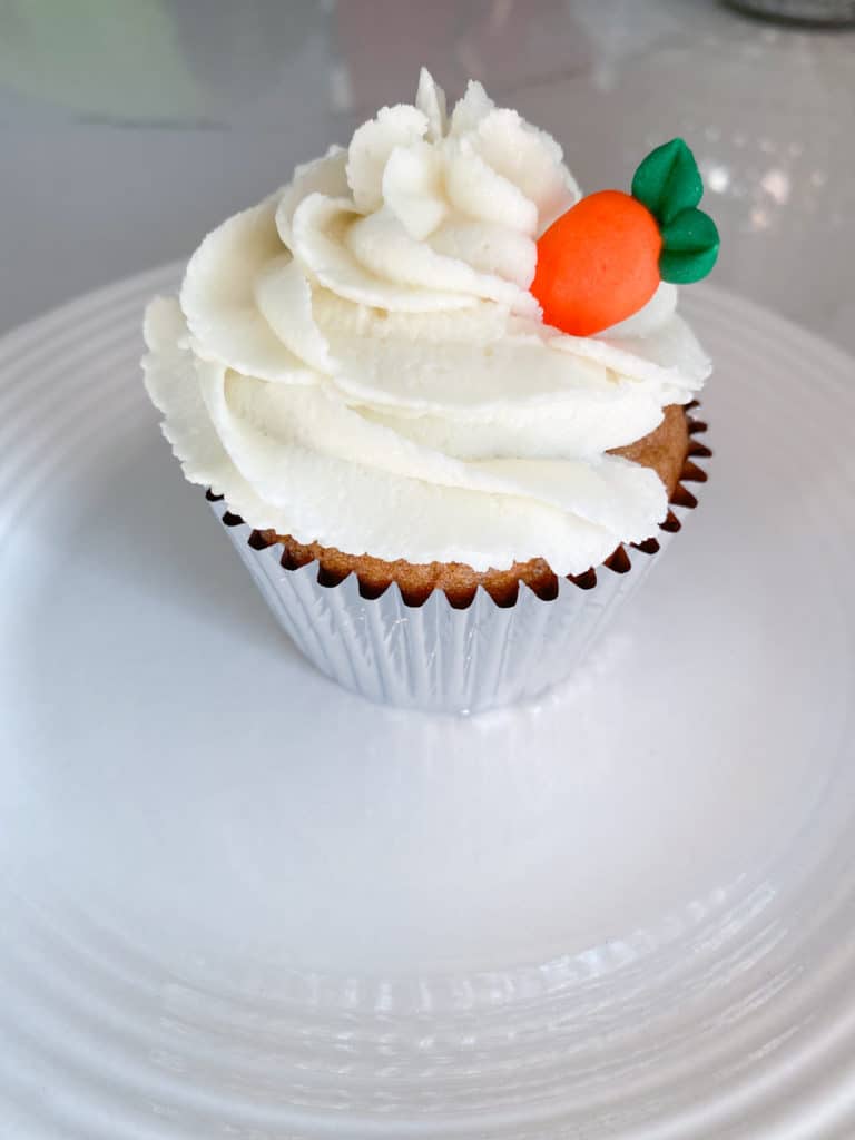 Cupcake in silver foil baking liner with ivory ginger buttercream and a royal icing carrot on a white plate