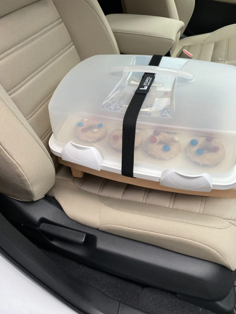 Stupid car tray with cupcake carrier strapped on top, on the front seat of a vehicle.