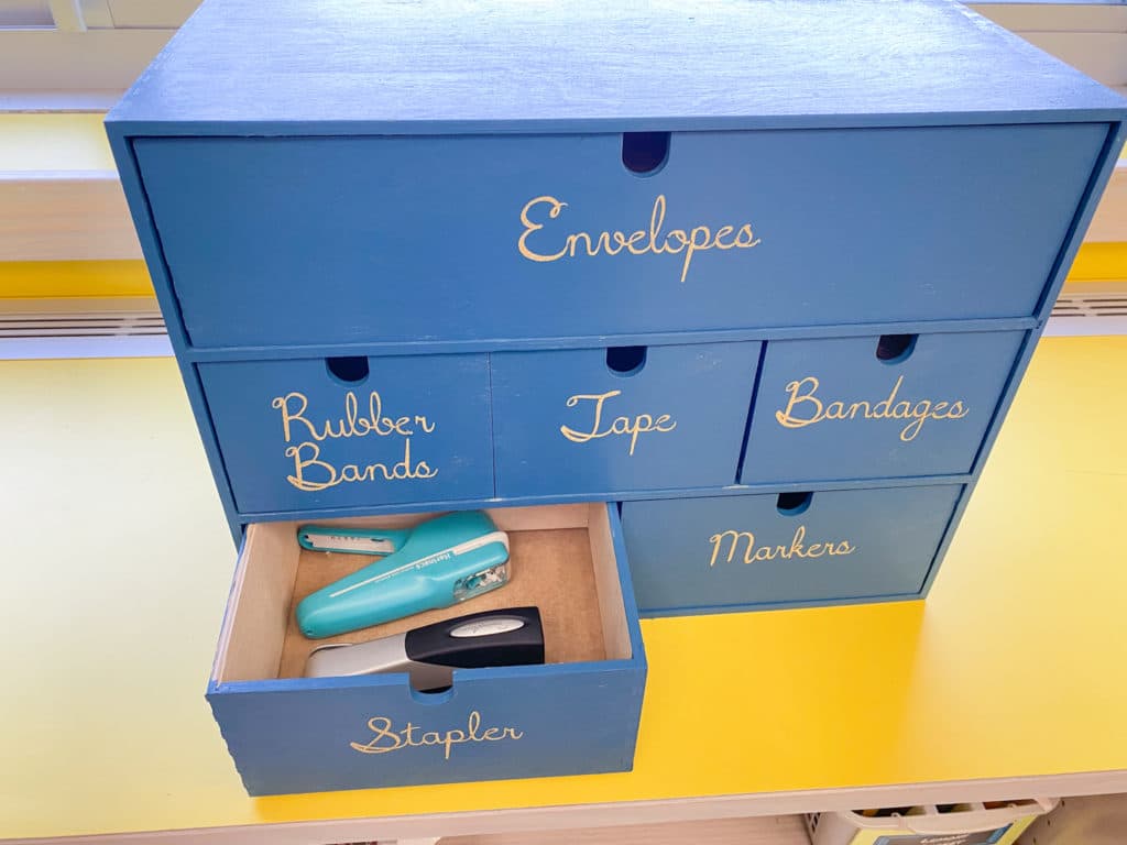 Blue teacher toolbox with labeled drawers for envelopes, rubber bands, tape, bandages, staplers and markers with 1 drawer open showing stapler and stapleless stapler from list of school supplies for teachers