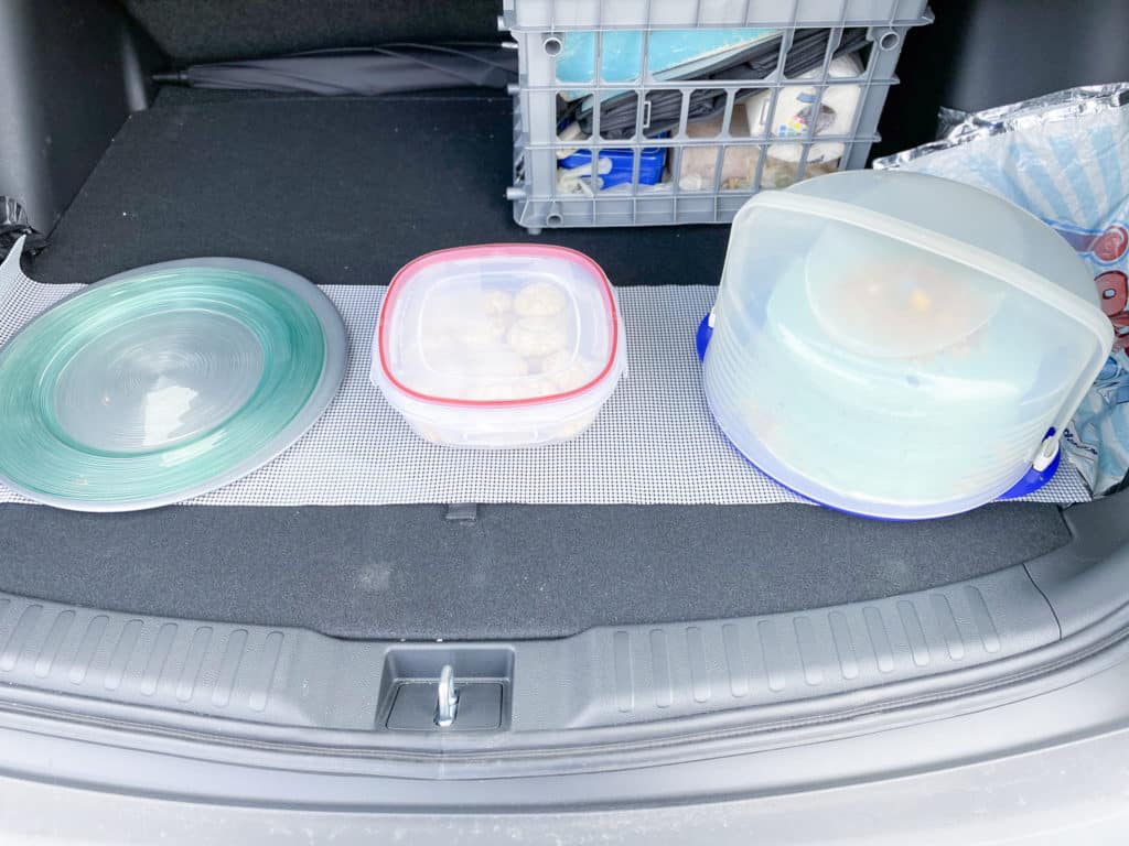 light gray Silicone shelf liner rolled out on floor of SUV trunk with metal charger dish with glass plate on top, square plastic storage container, and round plastic cake carrier  