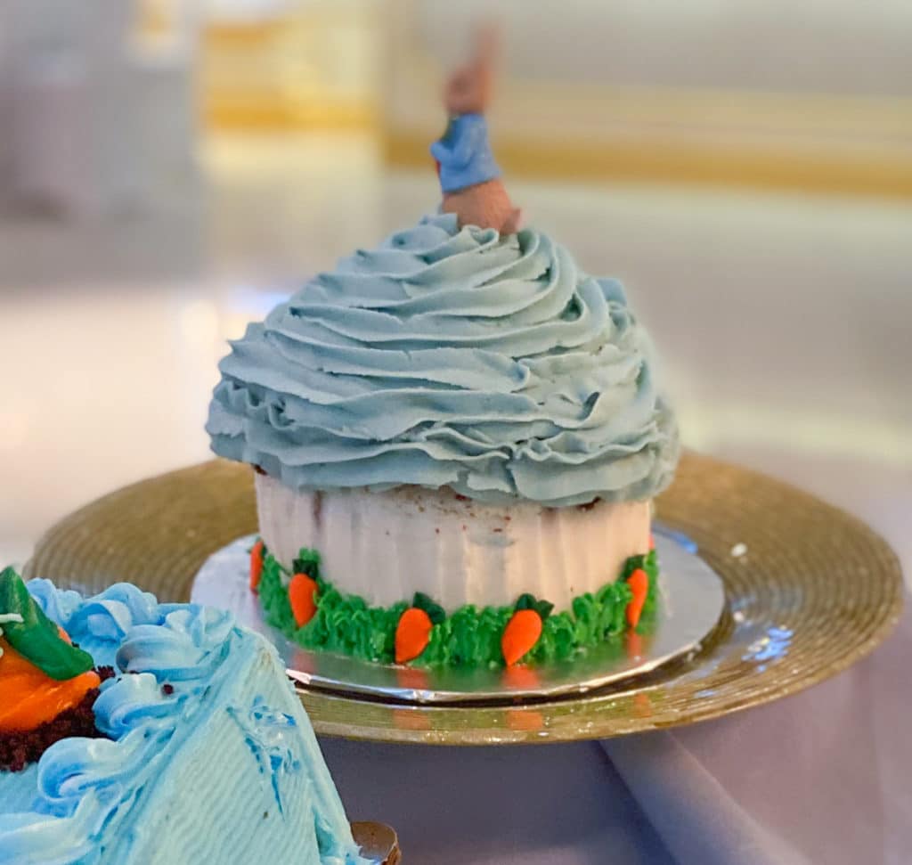 Giant cupcake smash cake with Peter Rabbit figurine on top, light blue icing, white buttercream cupcake liner, green grass and royal icing carrots on gold cake platter