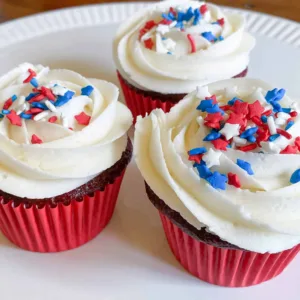 3 red velvet cupcakes with white vanilla buttercream icing and red, white and blue sprinkles on white platter