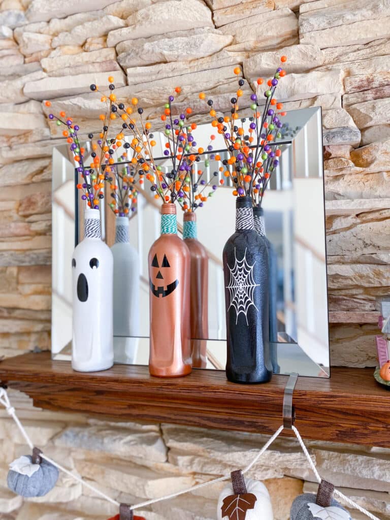 Halloween wine bottle craft with white ghost bottle, orange jack o'lantern bottle, and black bottle with white spider web design, with black and white twine and green and white twine around bottle necks and sprays of orange, black, purple and green berries in front of mirror on mantel