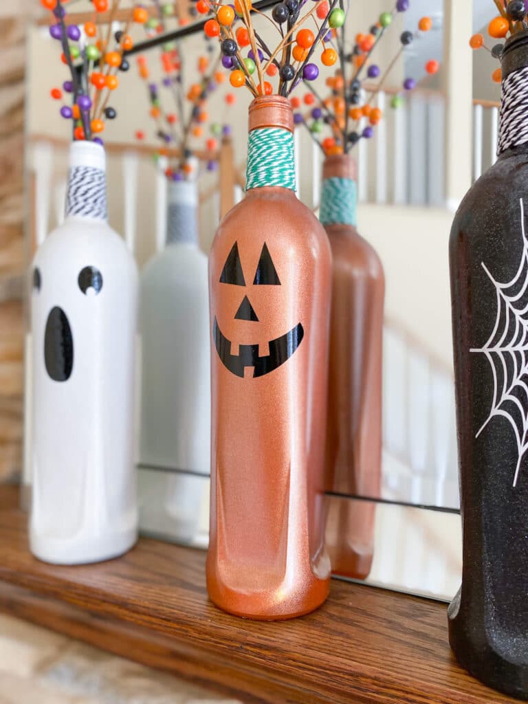 Cricut Halloween craft of Ghost face on white bottle, pumpkin face on orange bottle and white spider web on black bottle, in front of mirror on mantel
