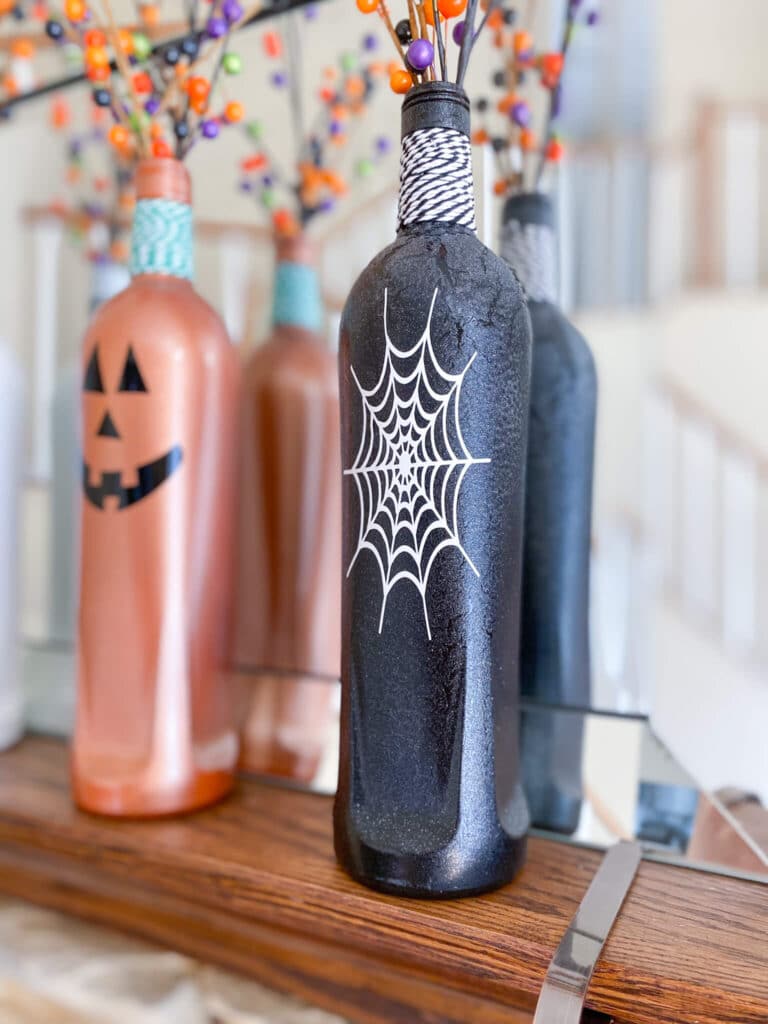 White spider web decal on black glitter bottle with streaky paint, next to orange jack o lantern bottle