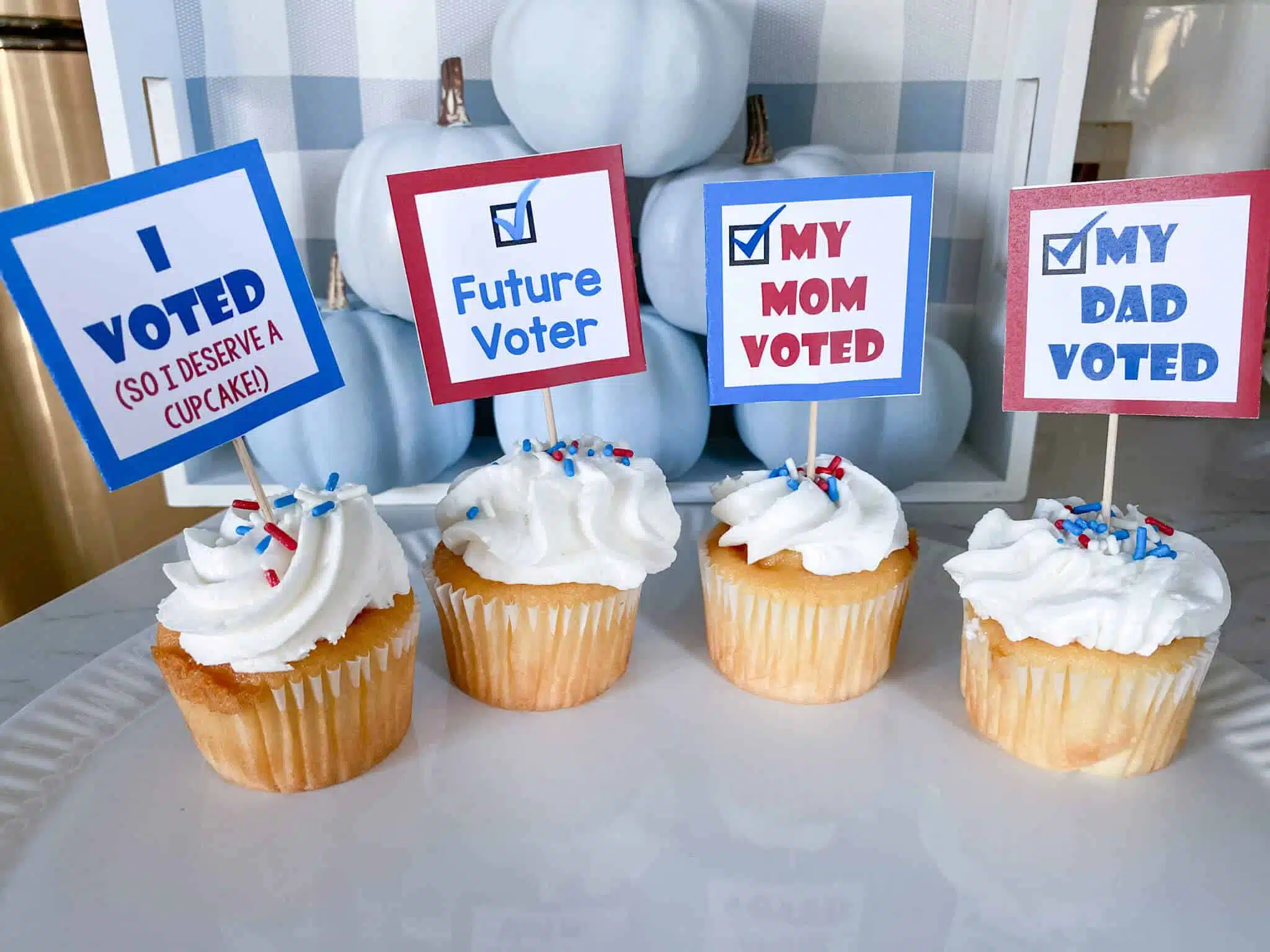 4 vanilla cupcakes with white frosting and red/white/blue sprinkles with square cupcake toppers on toothpicks: I Voted (so I deserve a cupcake), Future Voter, My Mom Voted, My Dad Voted, on white platter in front of blue ombre pumpkins
