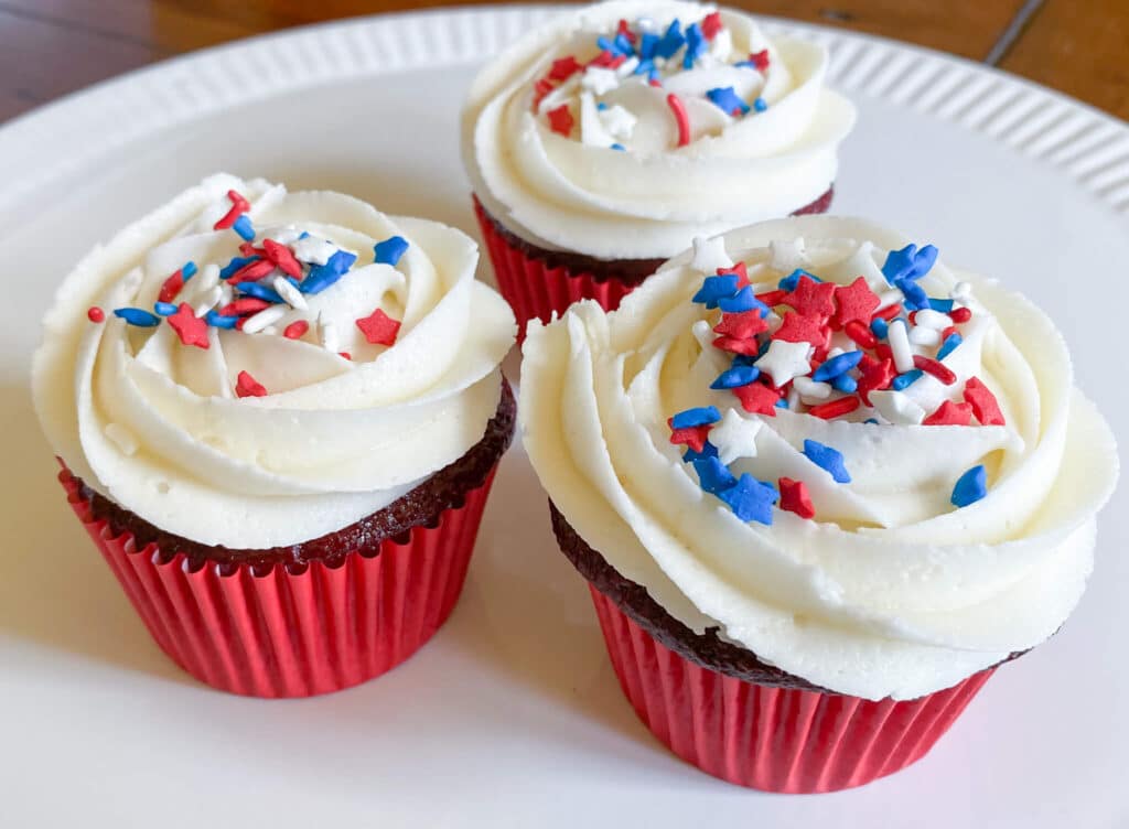 Red velvet cupcakes with vanilla icing and red, white and blue sprinkles with red cupcake wrappers on white platter
