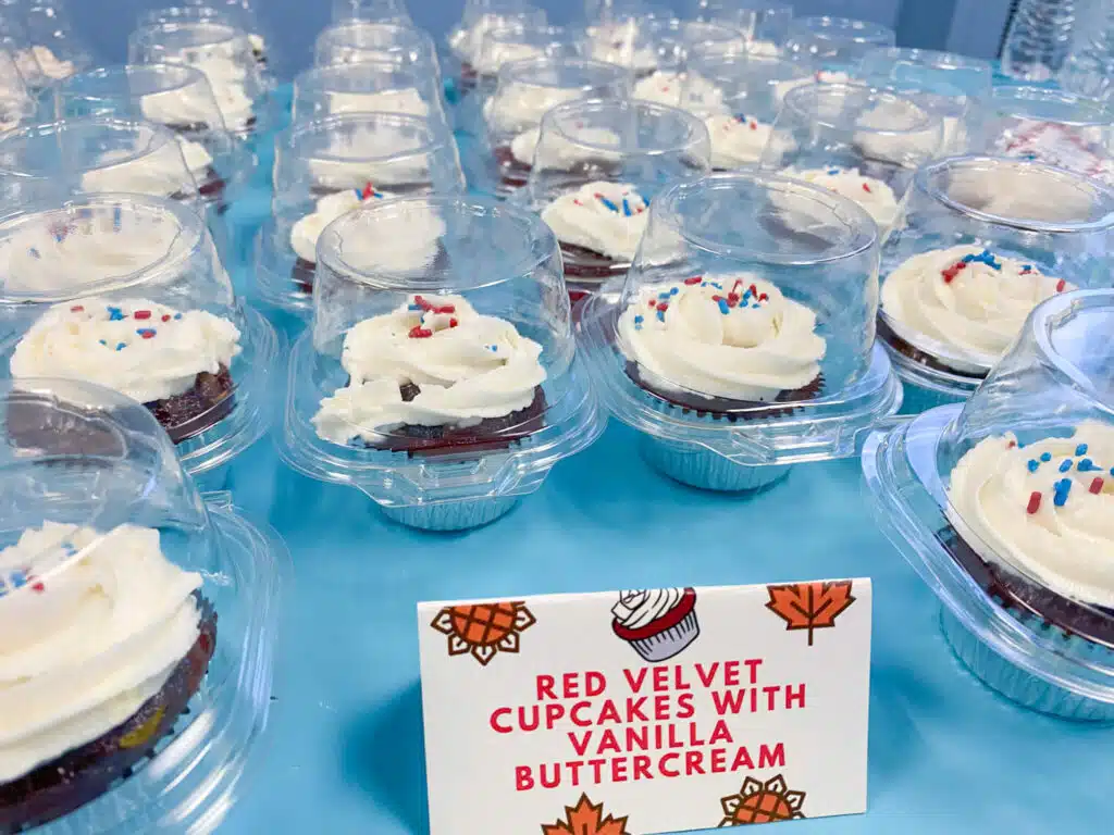 Rows of Red velvet cupcakes with vanilla swirl frosting and red/white/blue sprinkles in individual clear containers and sign that says Red velvet cupcakes with vanilla buttercream