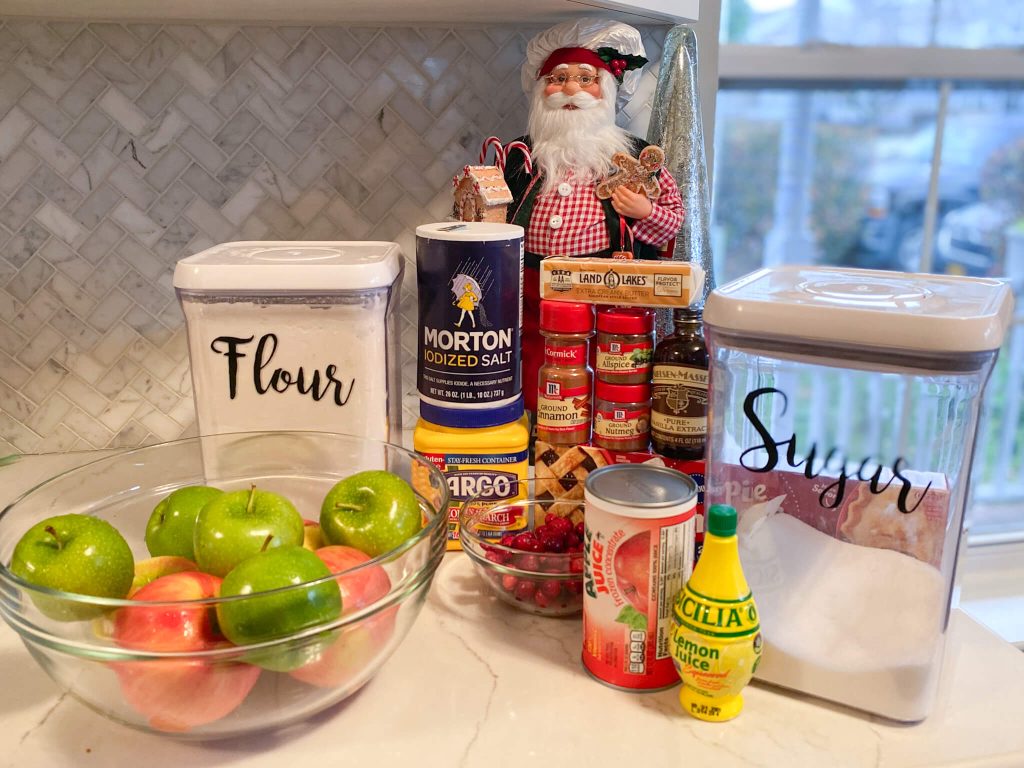 flour, sugar, salt, cornstarch, glass bowl of red and green apples, cinnamon, nutmeg, allspice, butter, vanilla extract, apple juice concentrate, lemon juice, cranberries, pie crusts in front of Santa Claus doll
