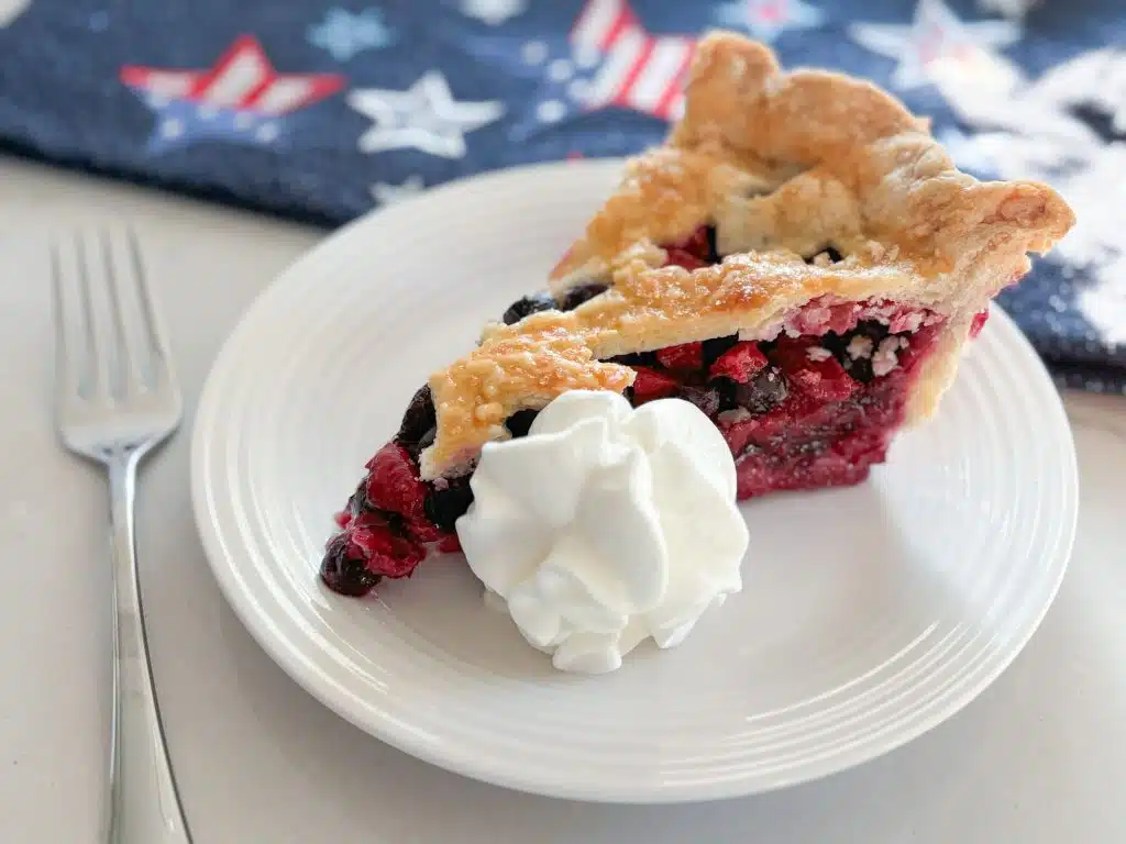 slice of strawberry blueberry pie with dollop of whipped cream on white plate in front of navy blue dish towel with red, white and blue stars