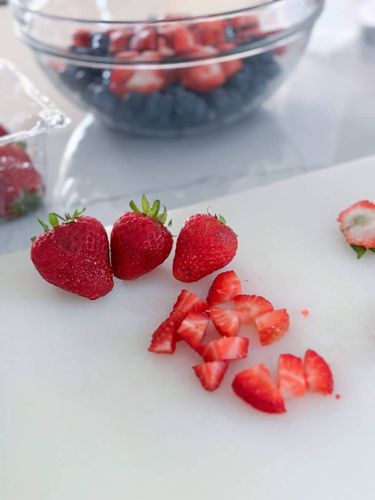 3 whole strawberries and one diced strawberry in front of glass bowl of strawberries and blueberrries