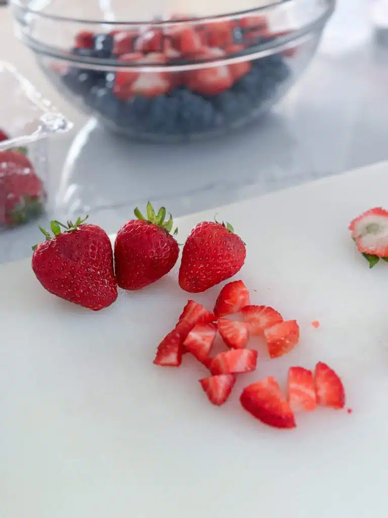 3 whole strawberries and one diced strawberry in front of glass bowl of strawberries and blueberrries