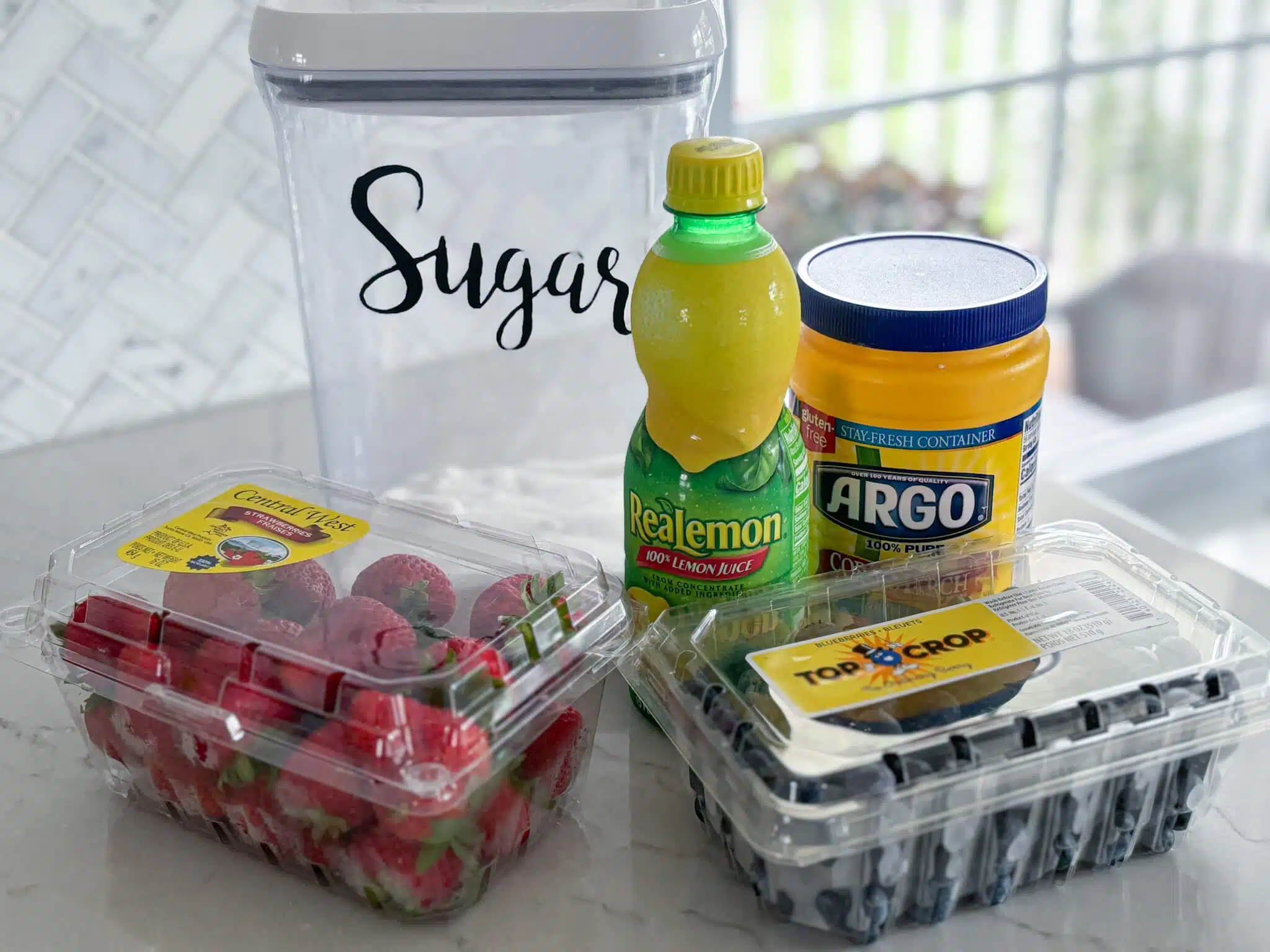 clear plastic container of strawberries, clear plastic container of blueberries, bottle of lemon juice, container of cornstarch, canister of sugar