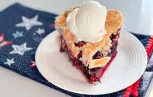 Slice of strawberry blueberry pie with scoop of vanilla ice cream on top on a white plate on top of a navy blue napkin with red, white and blue stars