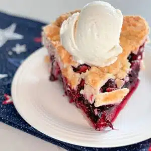 Slice of strawberry blueberry pie with scoop of vanilla ice cream on top on a white plate on top of a navy blue napkin with red, white and blue stars