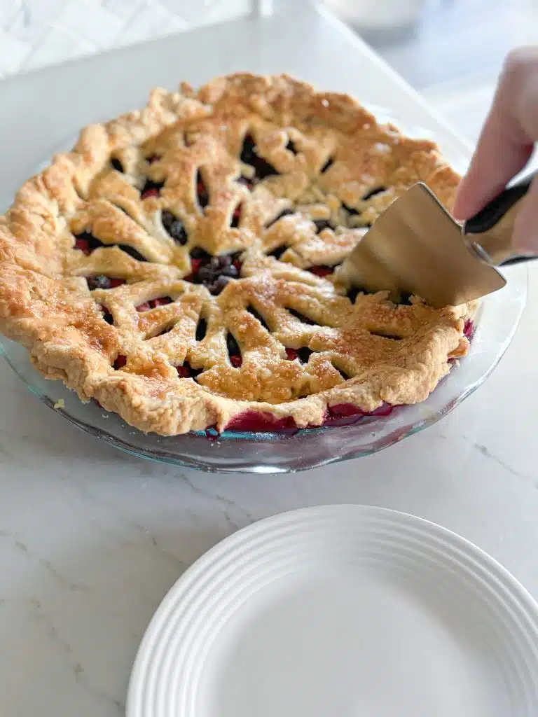 Pie spatula cutting into baked strawberry blueberry pie in front of white plate