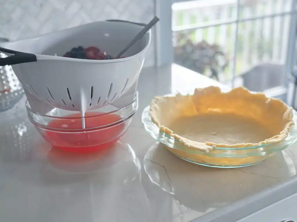 white colander with strawberries and blueberries draining red syrup into glass bowl next to glass pie plate of bottom crust