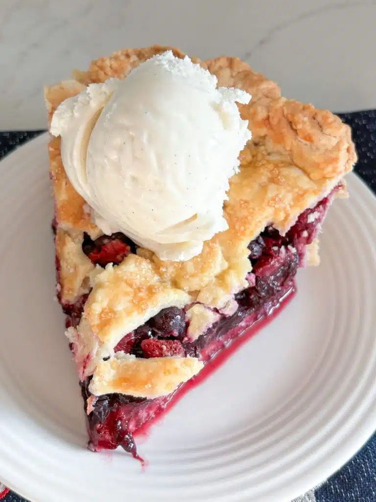 slice of strawberry blueberry pie with scoop of vanilla ice cream on white plate on top of blue dish towel