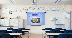 Classroom with desks in foreground facing whiteboard with blue slide that reads Welcome to Ms. Smith's and Mrs. Wagner's 5th Grade Class Back to School Night 2024, with alphabet above the board