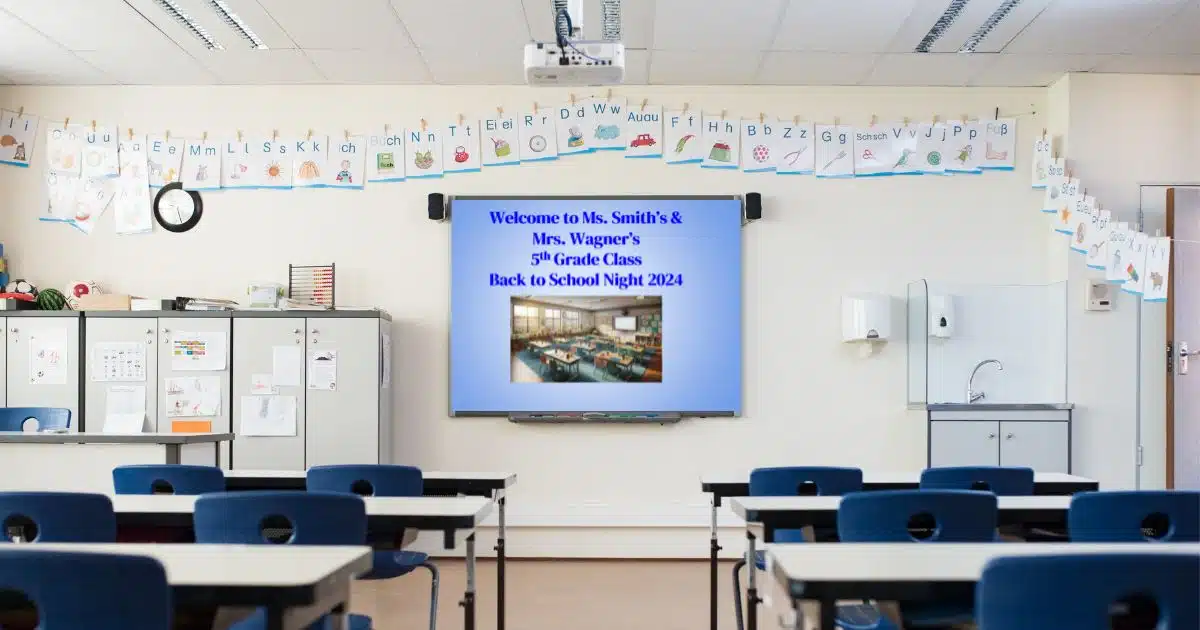 Classroom with desks in foreground facing whiteboard with blue slide that reads Welcome to Ms. Smith's and Mrs. Wagner's 5th Grade Class Back to School Night 2024, with alphabet above the board