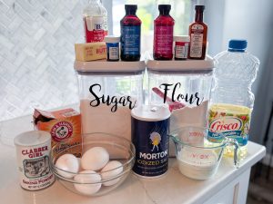 canisters of sugar and flour with bowl of eggs, salt, measuring cup of milk, baking soda, baking powder, butter, vegetable oil, vanilla extract, almond extract, blueberry emulsion, raspberry emulsion, red and blue food color gels