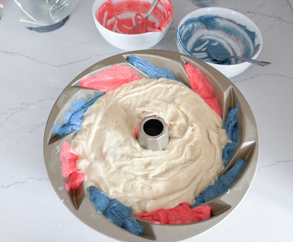 Heritage Bundt pan with white batter on top of red and blue swirls of batter, with bowls of red and blue batter behind it