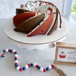 red white blue bundt cake wih marshmallow glaze and sprinkles on white cake stand with red white and blue beads in foreground