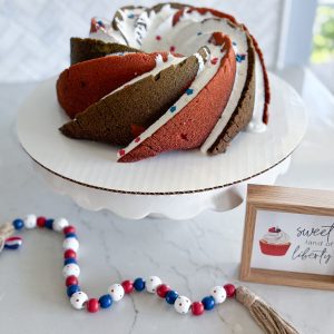red white blue bundt cake wih marshmallow glaze and sprinkles on white cake stand with red white and blue beads in foreground