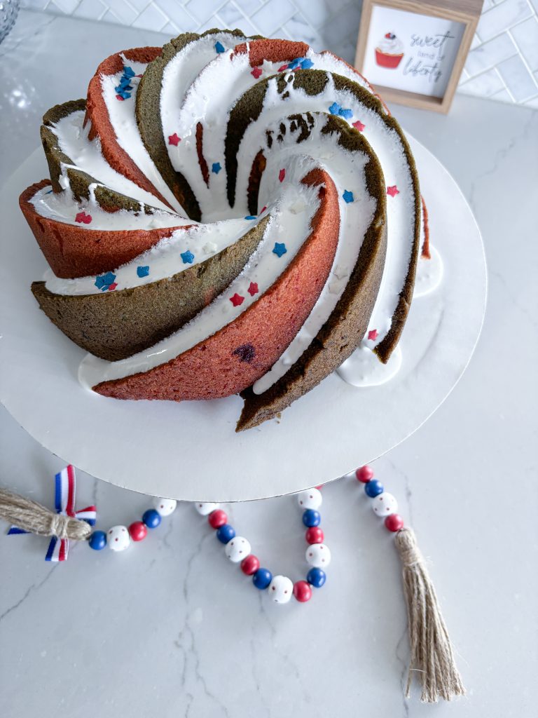 overhead view of red white and blue bundt cake wiht white glaze, red white and blue star sprinkles on white cake stand with red white and blue beads below it