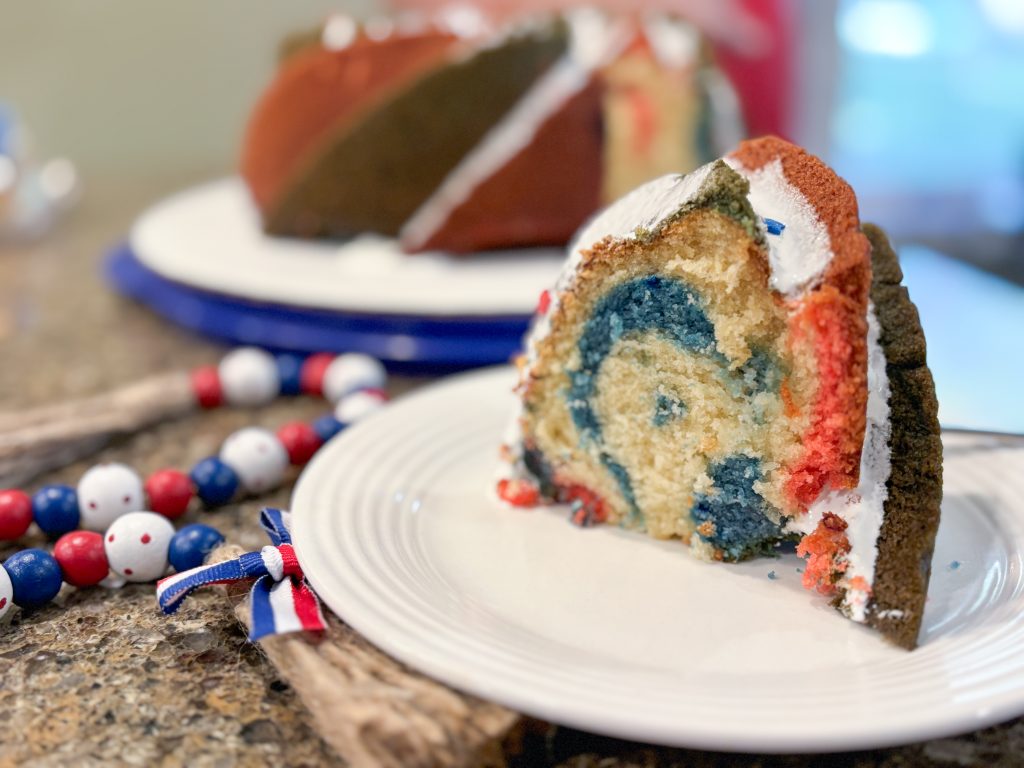 Slice of red white and blue bundt cake on white  plate in front of platter with rest of cake on it, red white and blue beads next to it