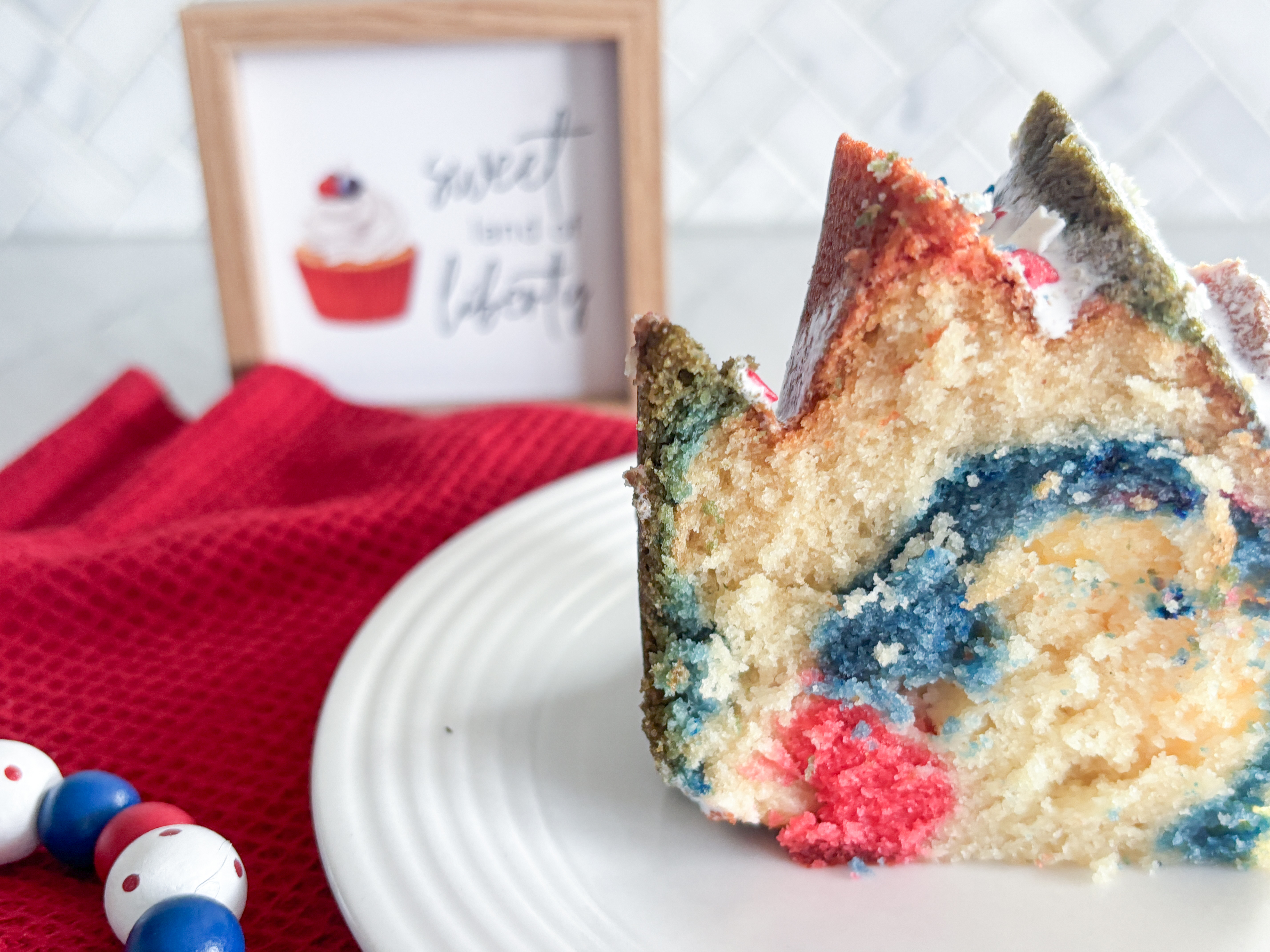 slice of bundt cake with red white and blue swirls on white plate, red napkin, red white and blue beads and sign that says sweet land of liberty