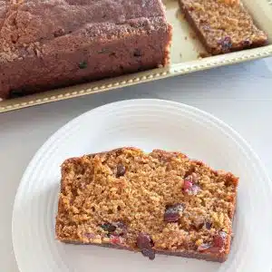 Slice of cranberry pumpkin bread on white plate next to loaf of pumpkin bread on gold rectangular platter