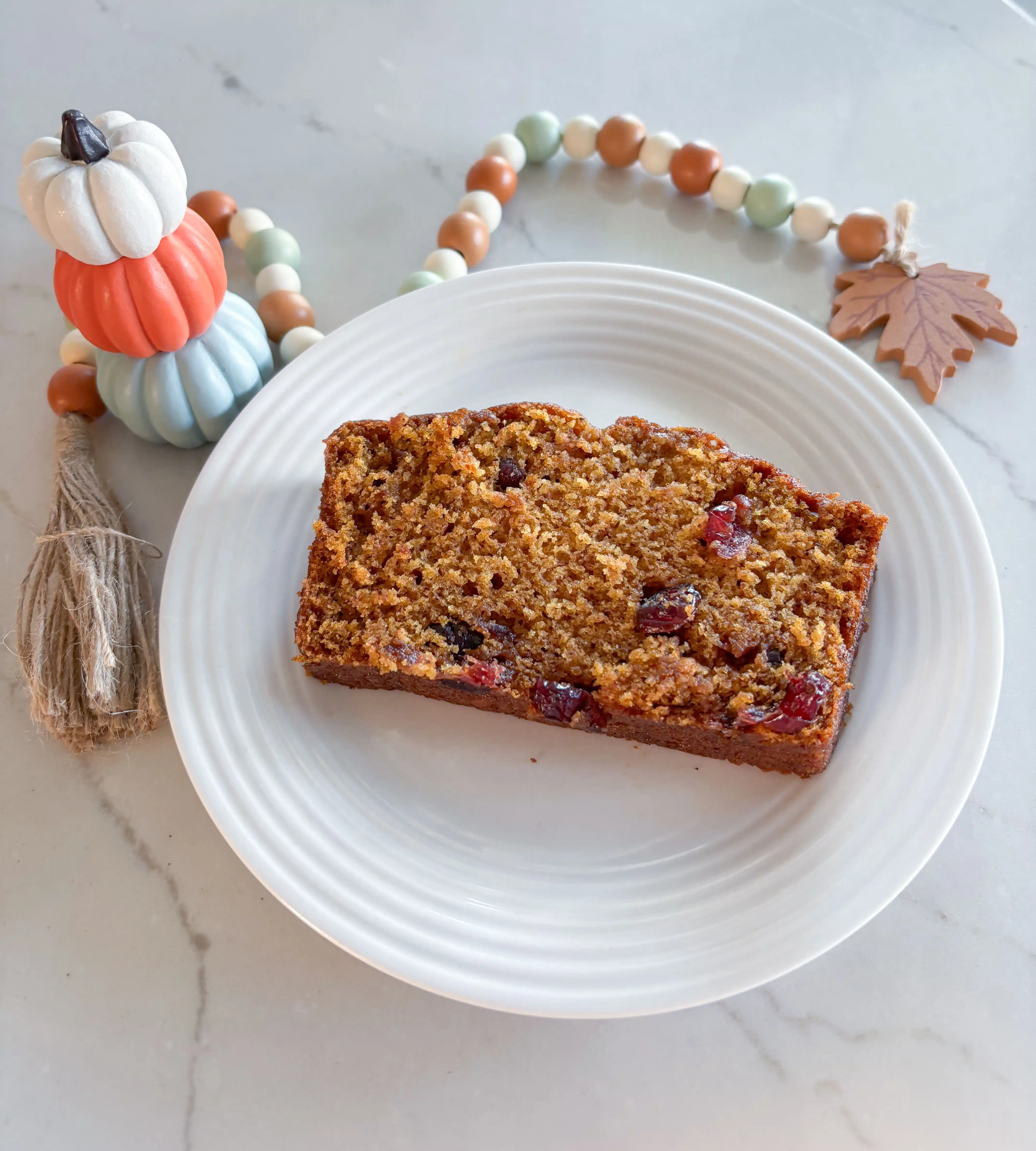 slice of cranberry pumpkin bread on white dish next to fall beads and stack of white/orange/light blue pumpkins