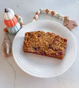 Slice of cranberry pumpkin bread on white dish next to figurine of white, orange and light blue pumpkins and wooden beads with fall leaf and tassel